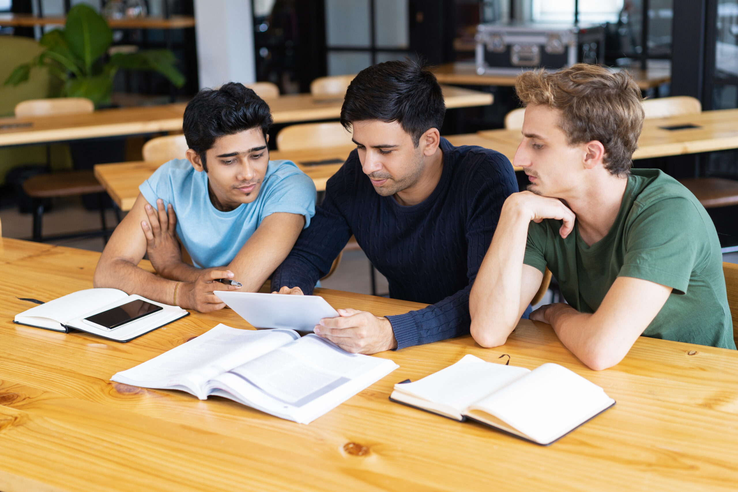 Three serious students studying and using tablet computer together. Young men sitting at desk in classroom or library. Education and technology concept.